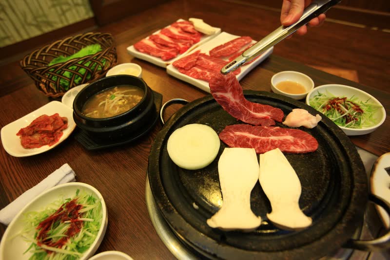Tongs lift a slice of red beef over a round black grill surrounded by soybean stew, salad and small side dishes