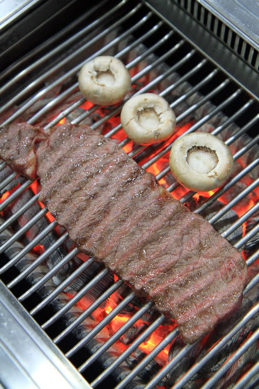 A long strip of marbled hanwoo beef searing on a grate over glowing charcoal, with mushrooms beside it