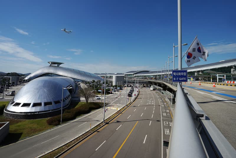 Roads and walkways outside the passenger terminal at Incheon International Airport under a blue sky, with a plane overhead and Korean flags on a pole