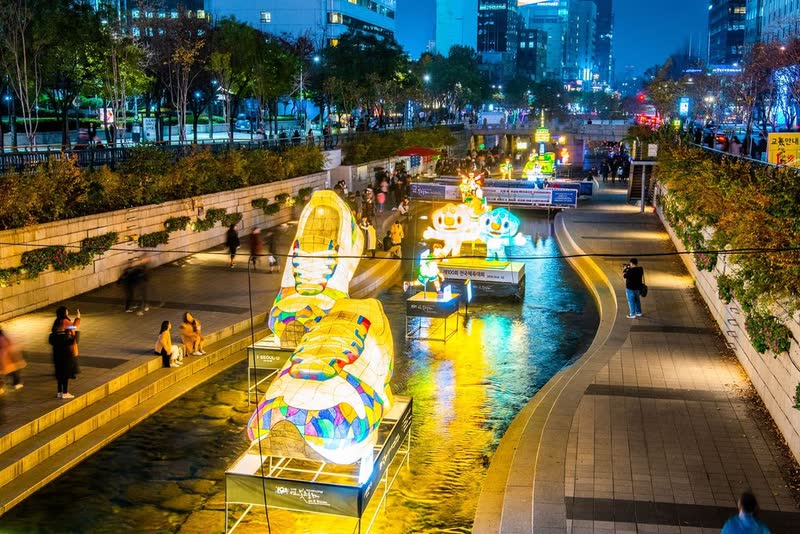 Glowing lantern sculptures standing in Cheonggyecheon Stream at night, with walkers on the paths and office towers above