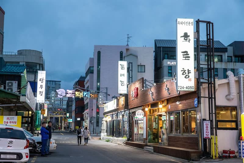 Jeju City's black pork street at dusk, a row of barbecue restaurants with lit signs