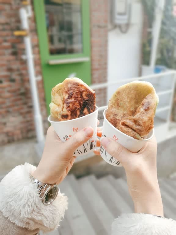 Two hands holding hotteok in paper cups on a cold day, one cut open to show dark syrup