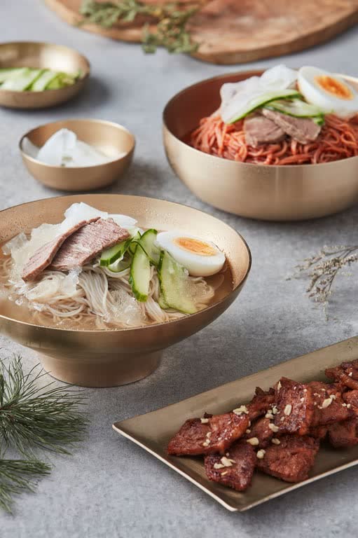 Two bowls of cold noodles, one in icy broth and one spicy, with a plate of grilled galbi