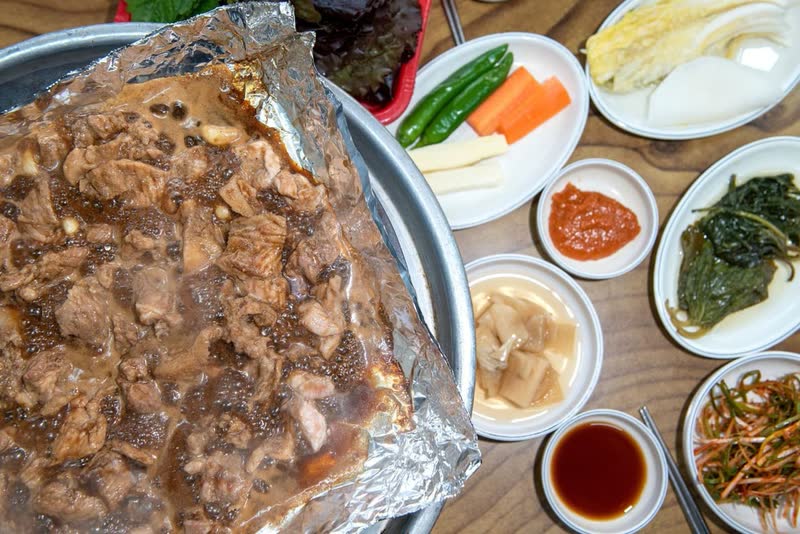 Marinated pork galbi on foil on a grill, surrounded by side dishes and dipping sauce