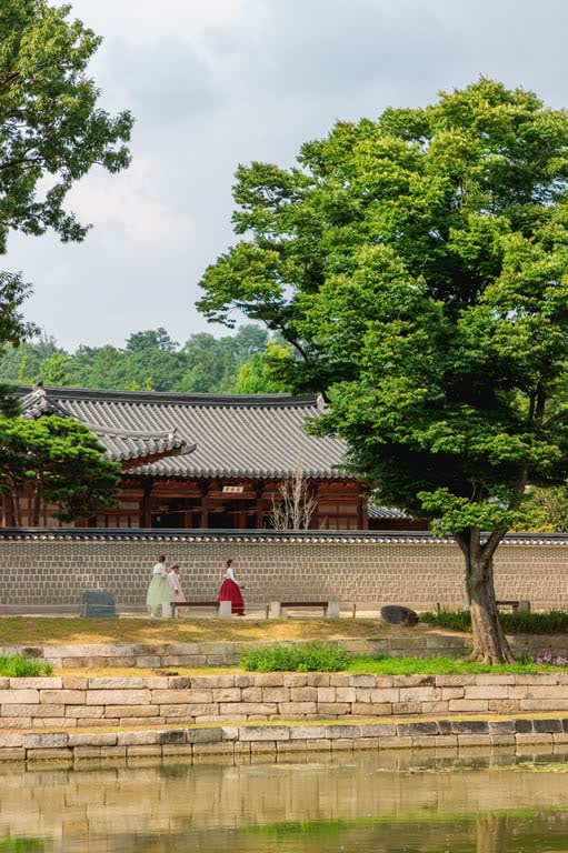 Small figures in hanbok walking along a long stone wall inside Gyeongbokgung, with a tiled-roof hall and a large tree above a pond