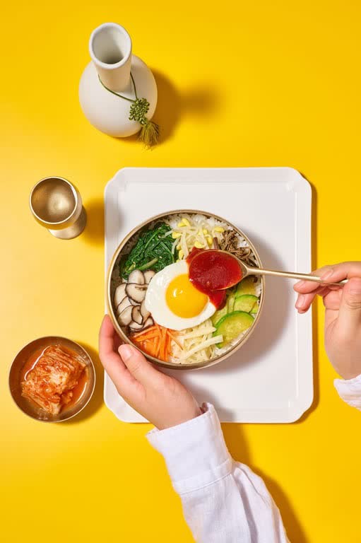 A bowl of bibimbap with seasoned vegetables, mushrooms and a fried egg, with a spoon adding red chili paste, kimchi on the side