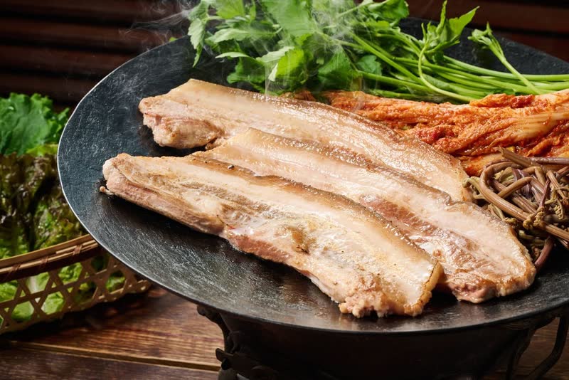 Thick slices of golden grilled pork belly on a round black cast-iron lid, with kimchi and greens at the edge
