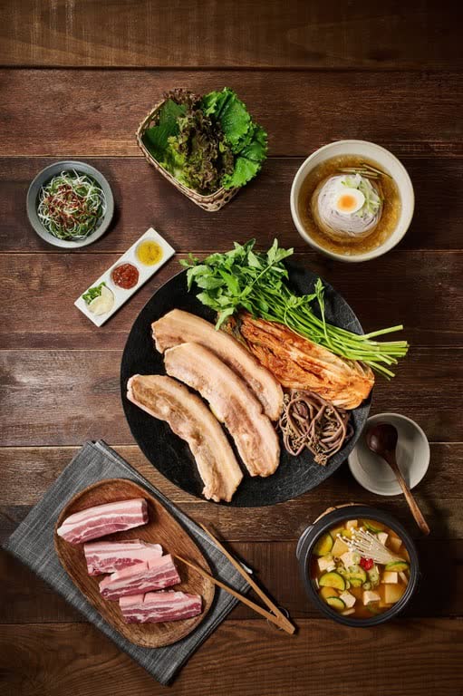 A samgyeopsal table from above: grilled pork belly on a round iron pan, raw belly on a plate, lettuce, dipping sauces and a bowl of soybean stew
