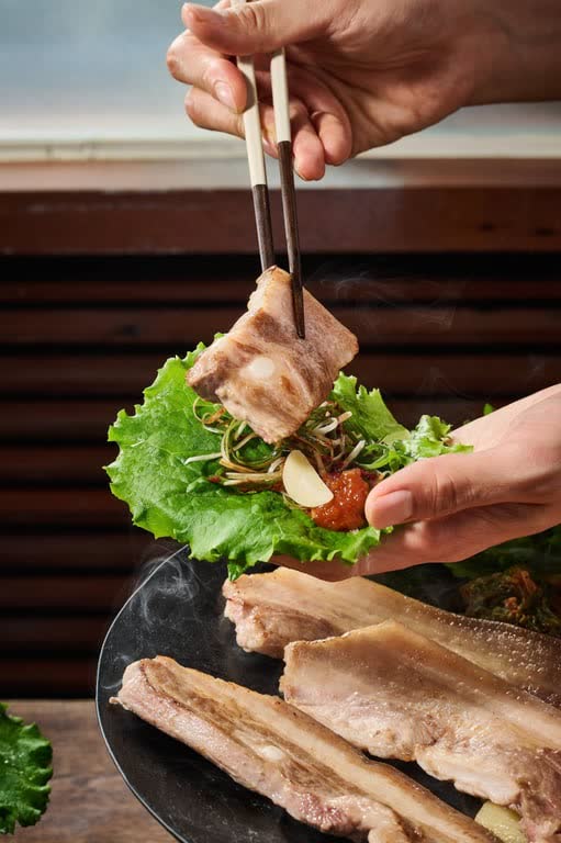 Hands holding a lettuce wrap with a piece of grilled pork belly and garlic, above a pan of grilled belly