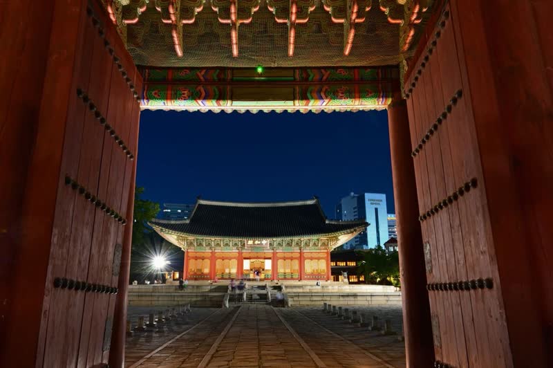 A lit palace hall at Deoksugung seen at night through an open wooden gate