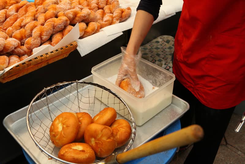A vendor in gloves rolling fresh round doughnuts in a tub of sugar, with twisted doughnuts piled behind