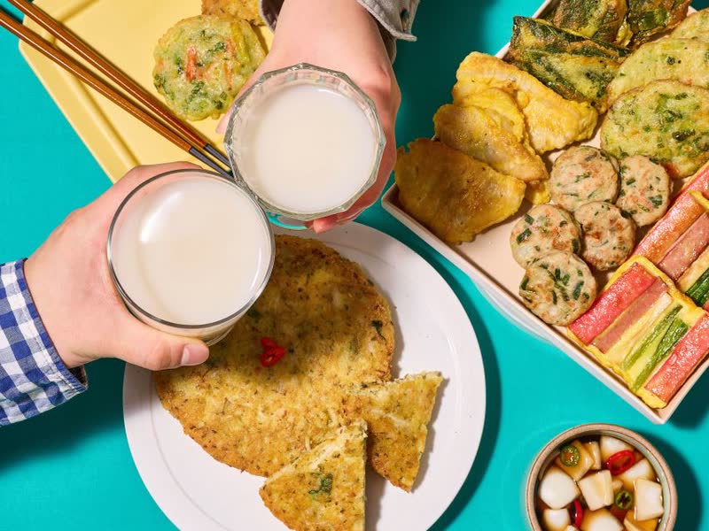 A table of assorted jeon (savory pancakes) with two glasses of milky makgeolli raised in a toast