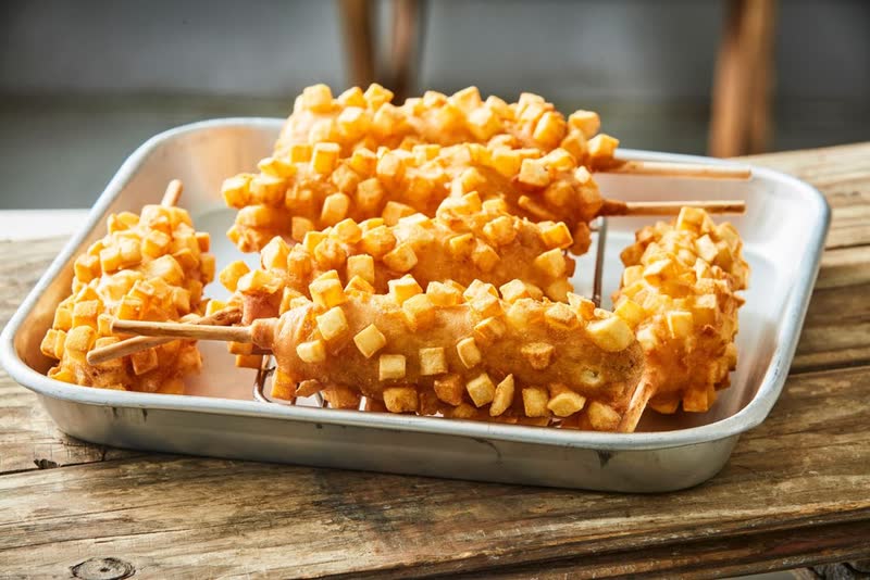 Korean corn dogs coated in cubes of fried potato, on sticks in a metal tray