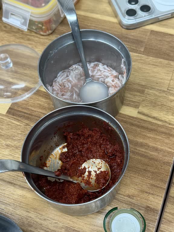 Two steel tins at the side of a table, one of salted tiny shrimp and one of red seasoning paste, each with its own spoon