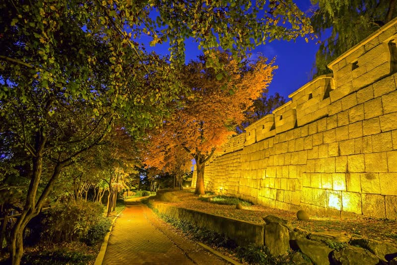 The Seoul City Wall at night along the Naksan trail, lit by warm lamps under autumn trees