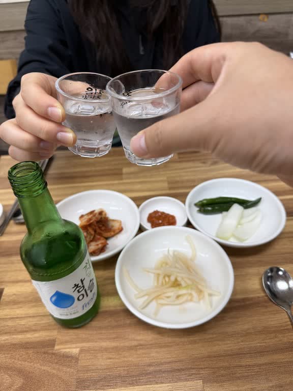 Two small glasses of soju clinking over a table of kimchi, pickled radish, raw green chilies and onion
