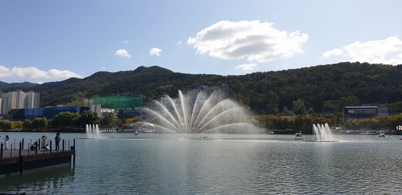 Fountain spraying over Suseongmot lake in Daegu on a sunny day, with a wooded hill and small boats behind